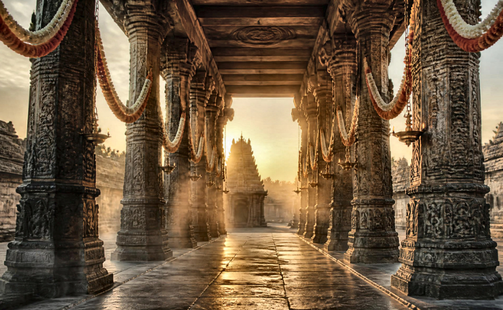 Sunlit stone corridor of a South Indian temple courtyard, carved pillars hung with garlands and brass lamps, leading toward a distant gopuram